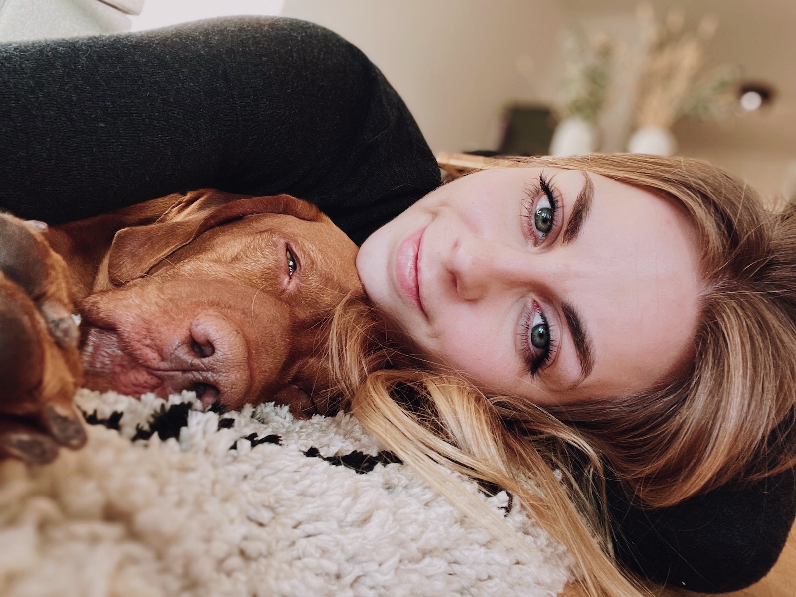 Junge Blonde Frau liegt mit mit einem großen braunen Hund entspannt im Arm kuschelnd auf einem schwarz-weißen Fransenteppich