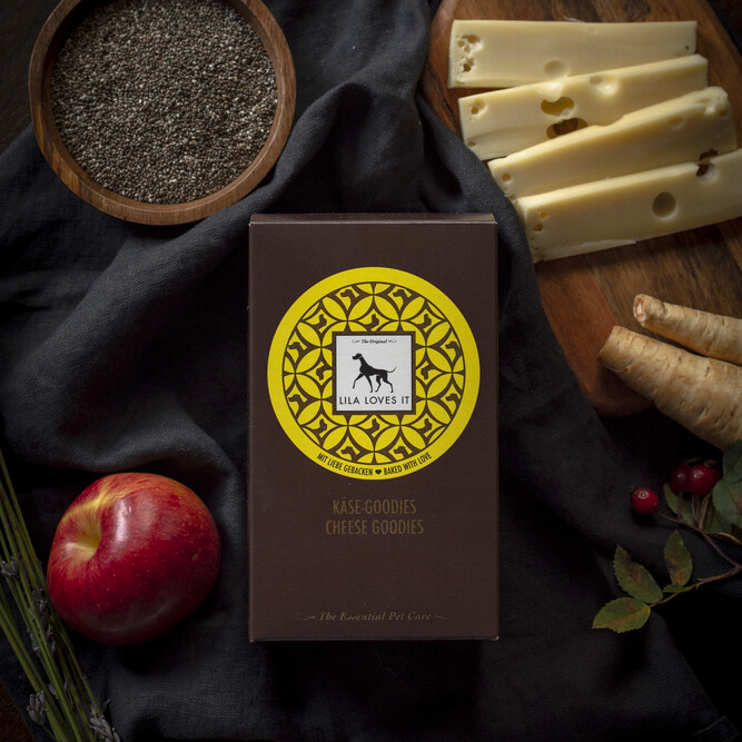 Eine elegante gelbe und braune Faltschachtel mit Leckerli für Hunde aus Käse liegt auf einer dunklen Decke neben einem Brett mit Käsestücken, Pastinaken, einem Apfel, Chia-Samen und Beeren und einem | LILA LOVES IT "Käse-Goodies"